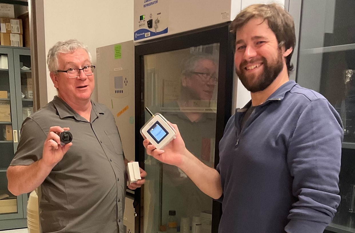 Smiling Christopher Holly and Ryan Davis holding monitoring units in front of a research freezer